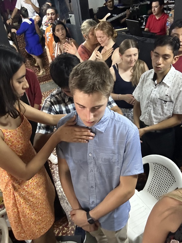 A girl wearing an orange dress praying over a boy wearing a blue collared shirt with other kids praying around them in a group